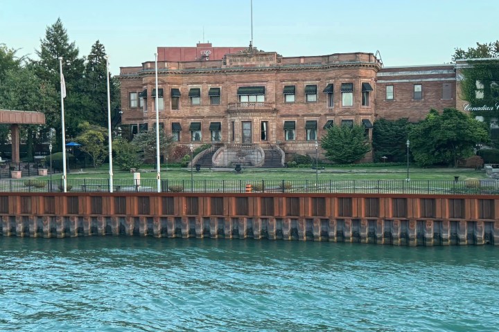 Historic brick building by water with trees and a lawn.